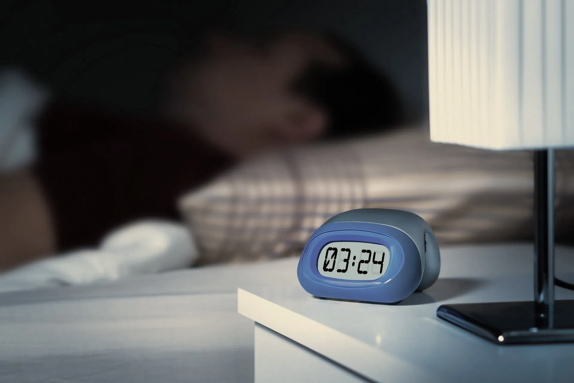 A bedside clock reads 3:24 a.m. next to a phone and a lamp, with a person in bed, out of focus, in the background.