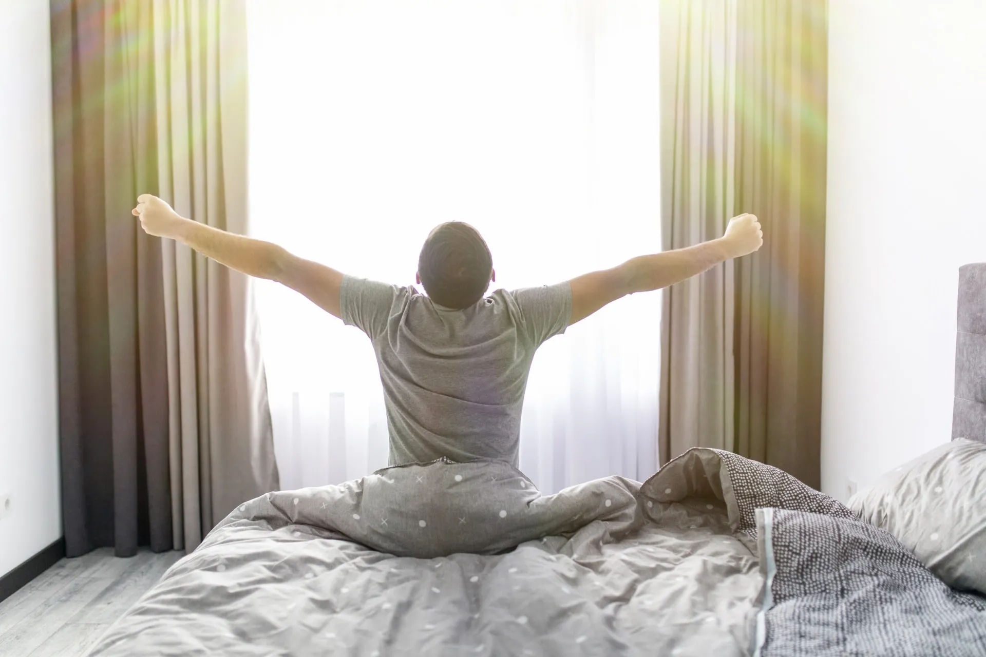 A man sits up in bed and stretches toward a bright window as morning light comes through the curtains